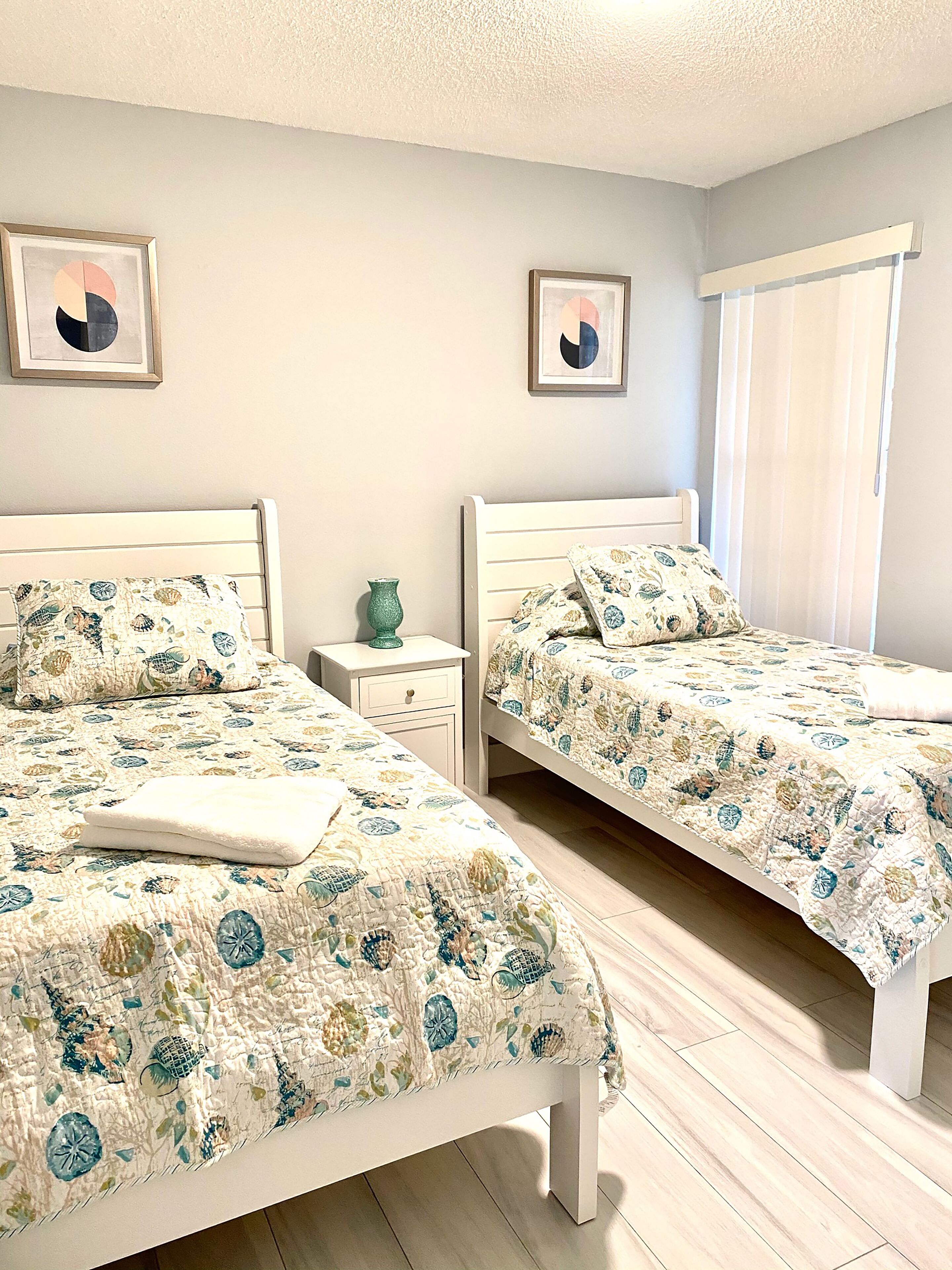 Un dormitorio con dos camas, cada una con ropa de cama floral a juego, una mesita de noche con un florero y una ventana con persianas.