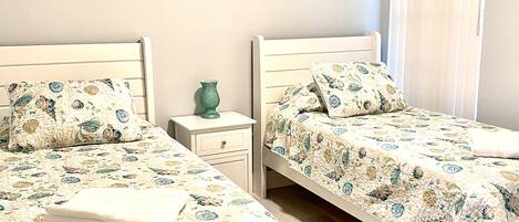 Une chambre à coucher avec deux lits, chacun agrémenté d’une parure de lit à motifs floraux assortis, une table de chevet avec un vase et une fenêtre équipée de stores.
