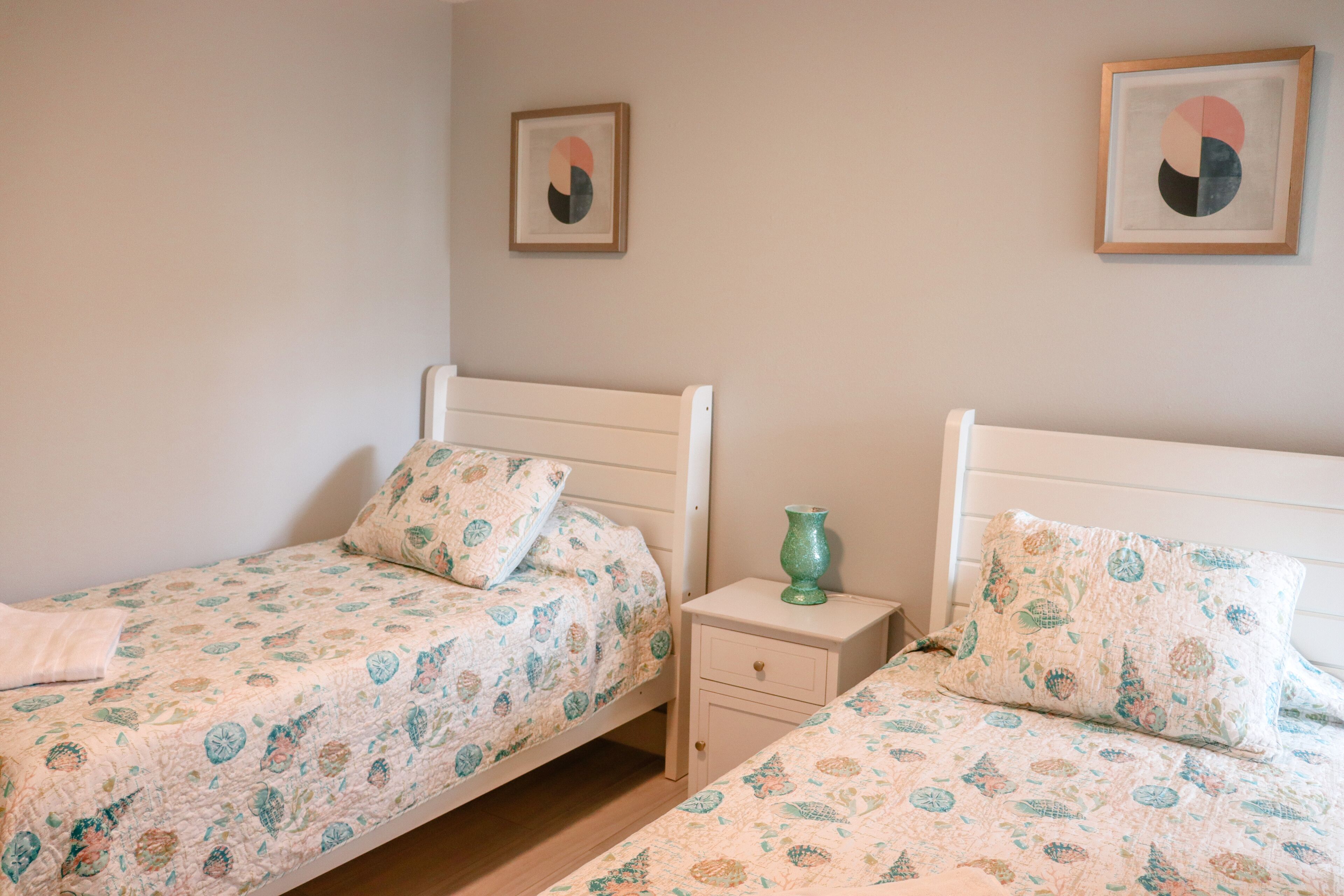 Une chambre avec deux lits simples, chacun agrémenté d’une parure de lit assortie à motifs floraux. Une petite table de chevet se trouve d’un côté, sur laquelle est posé un vase vert.