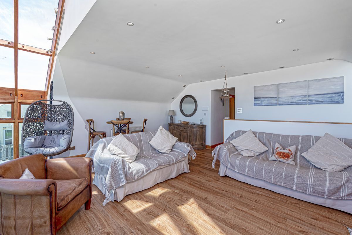A spacious living area with a high ceiling, wooden flooring, and large windows allowing natural light. There are two sofas, a chair, and a small table with decorative items.