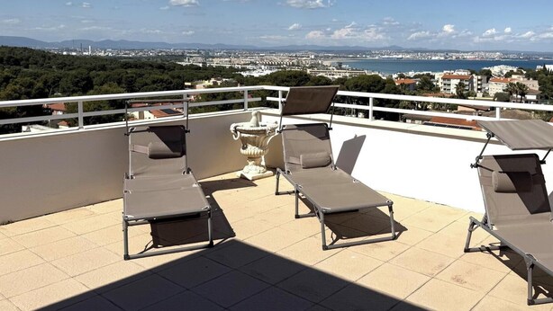 Eine Dachterrasse mit Liegestühlen, einem weißen Geländer und Blick auf eine Küstenstadt.