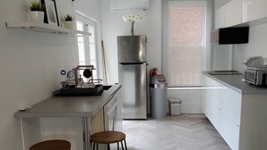 A modern kitchen with white cabinetry, a stainless steel refrigerator, a microwave, and a sink. There are two wooden stools and a small table with a plant. - Beautiful 2BR Suite just off Main St, Picton, near Sandbanks (Prince Edward)