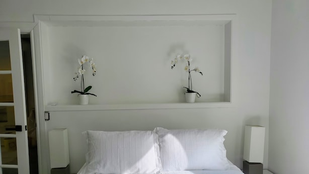 A neatly made bed with white linens, two white nightstands with lamps, and two white orchid plants on a shelf above the bed. - Beautiful 2BR Suite just off Main St, Picton, near Sandbanks (Prince Edward)