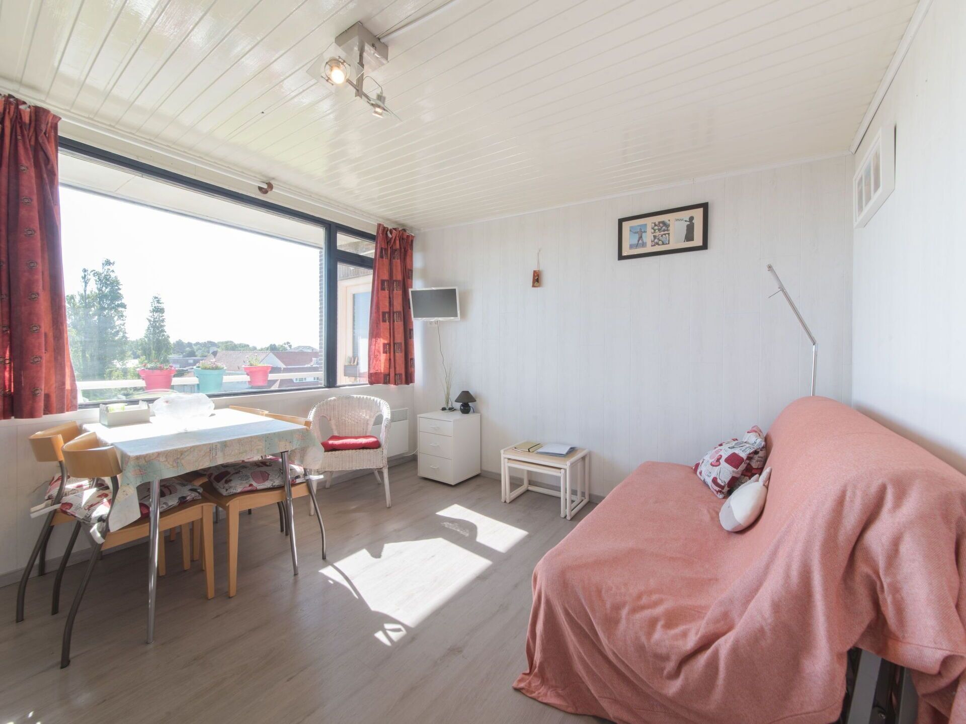 A room with a bed, a dining table with chairs, a small white cabinet, and a window with curtains.