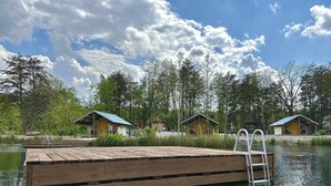 Un muelle de madera se extiende sobre un lago con una escalera, rodeado de cabañas con techos azules y un cielo despejado.