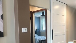 Ein Flur, der zu einem Schlafzimmer führt, mit einem Bett, einem Deckenventilator und einem Fenster mit Jalousien.