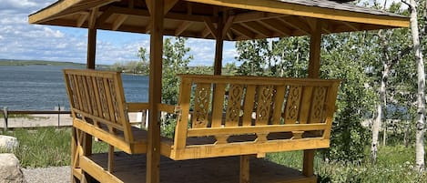 A wooden gazebo with a bench, located near a body of water with trees in the background.