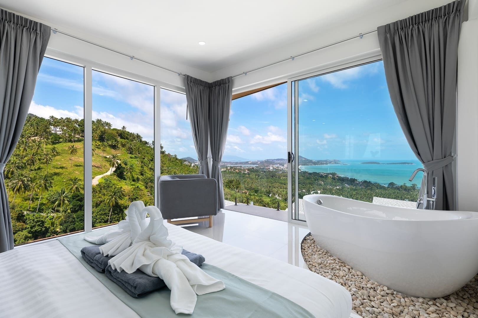 A modern bedroom with a large bed, a bathtub, and a view of a tropical landscape.