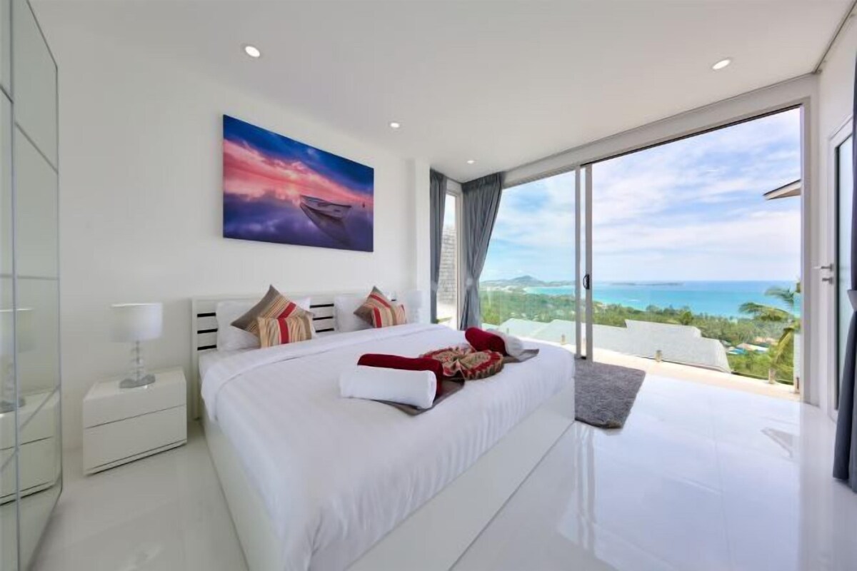 A modern bedroom with a large bed, a nightstand, a painting, and a view of the ocean.