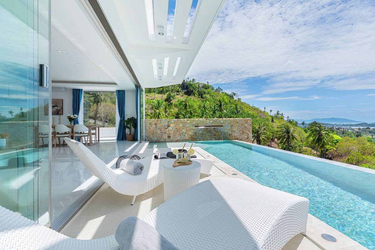 A modern outdoor pool area with white wicker furniture, a stone wall, and a view of a lush hillside.