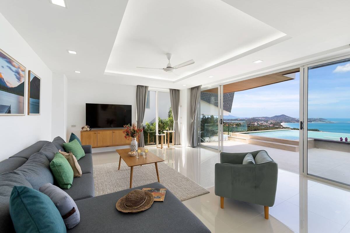 A modern living room with a large flat-screen TV, a sofa with green and gray cushions, a coffee table with decorative items, and a view of a pool and ocean.