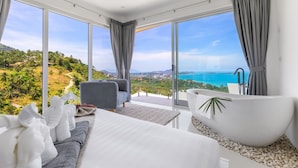 A modern bedroom with a large bed, a seating area, and a bathtub. It features large windows with a view of a coastal landscape. - Villa the wave (Koh Samui)