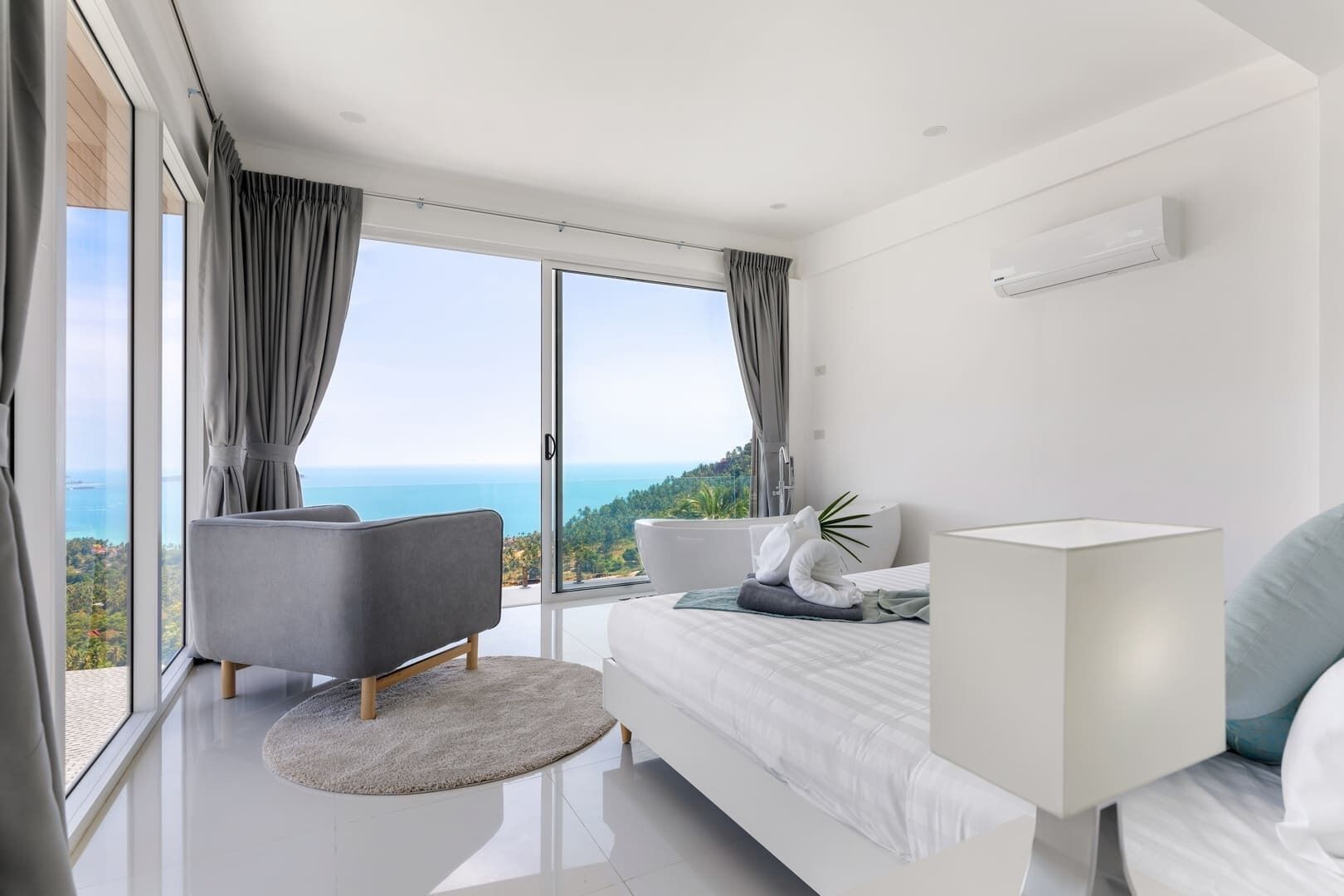 Une chambre à coucher moderne avec un grand lit, un fauteuil gris, un tapis rond et une vue sur l’océan.