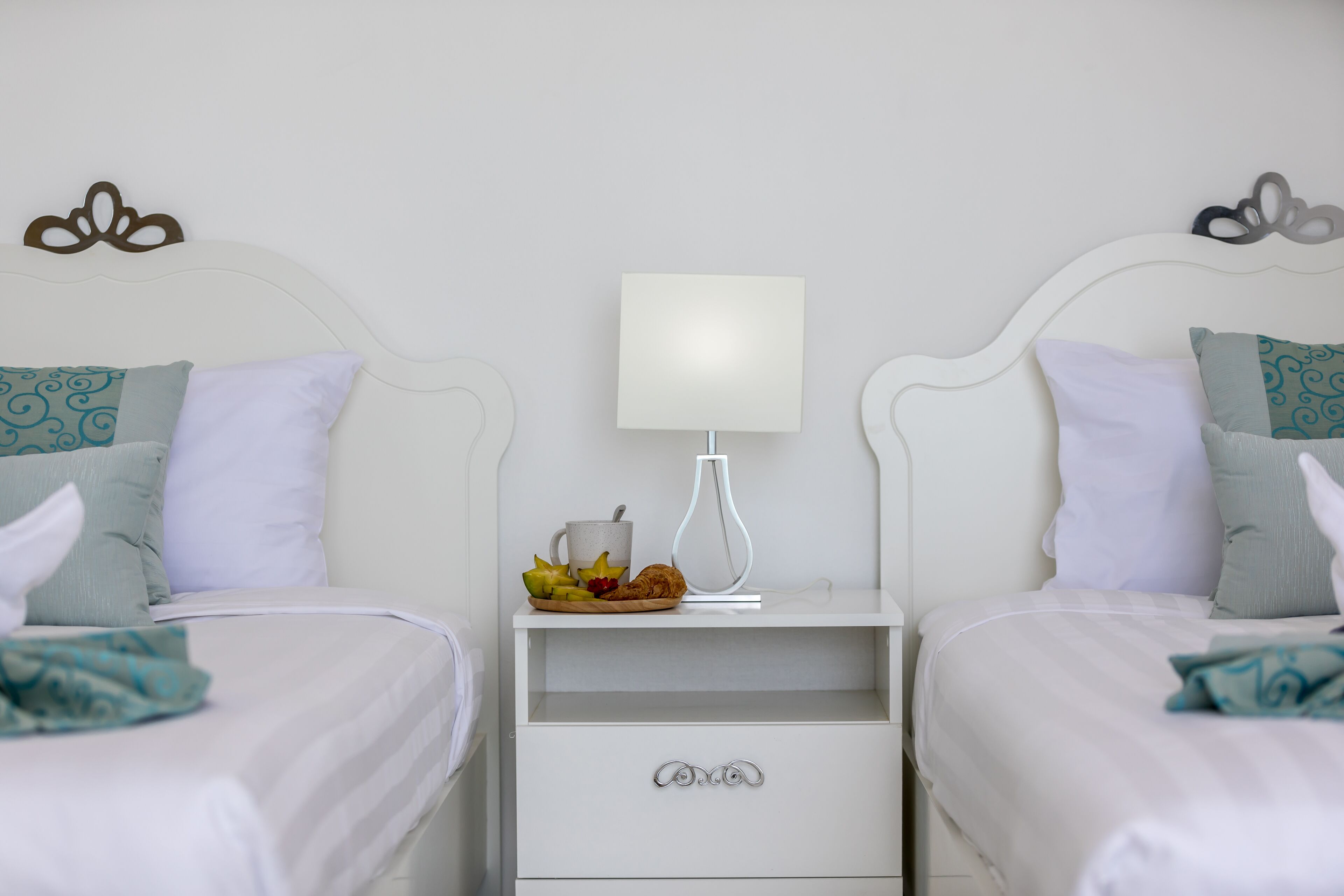 Et hotelværelse med to senge, et natbord med en lampe og et lille bord med frugt og en kop.