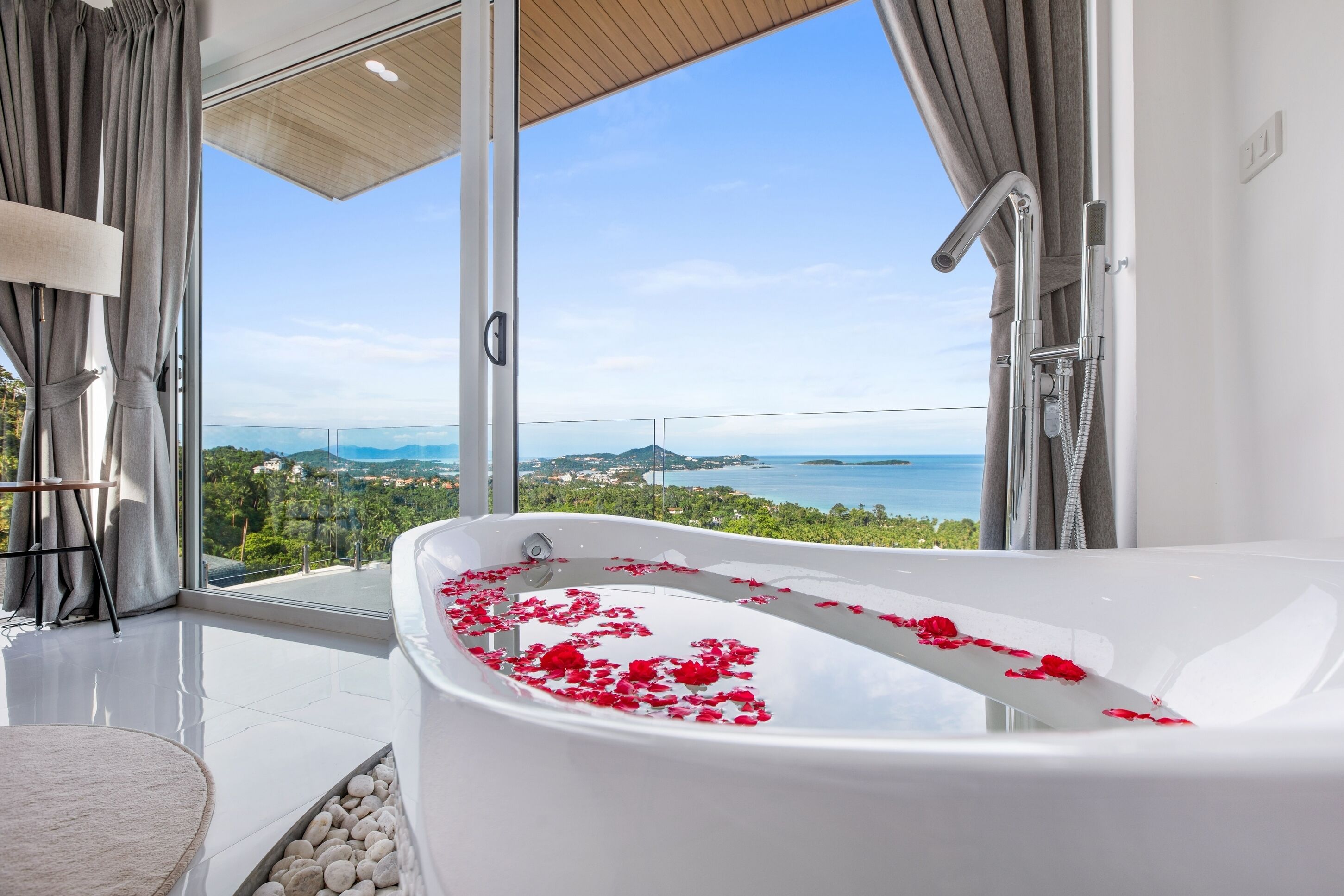 Ein modernes Badezimmer mit einer großen Badewanne, gefüllt mit Rosenblüten, Meerblick und einer gläsernen Duschkabine.