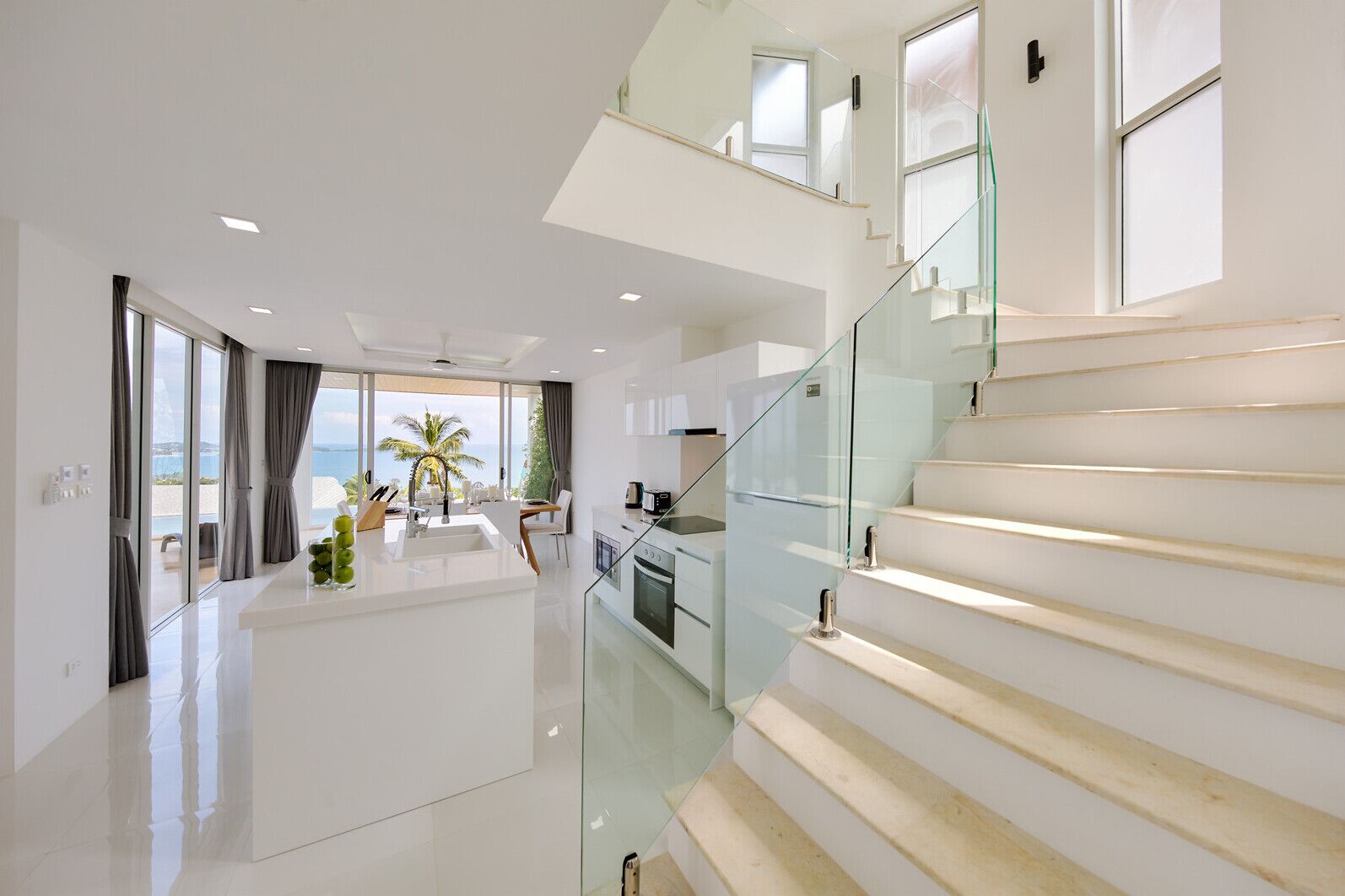 A modern interior with a glass staircase, a kitchen, and a dining area with a view of the sea.