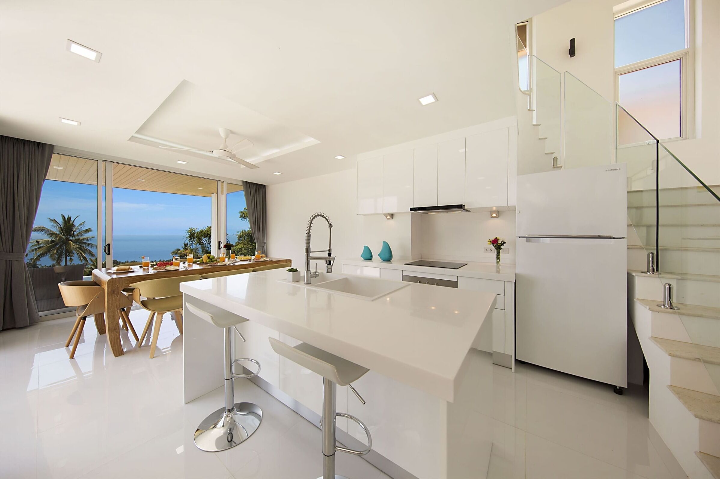 Uma cozinha moderna com ilha central, armários brancos e vista para o oceano.