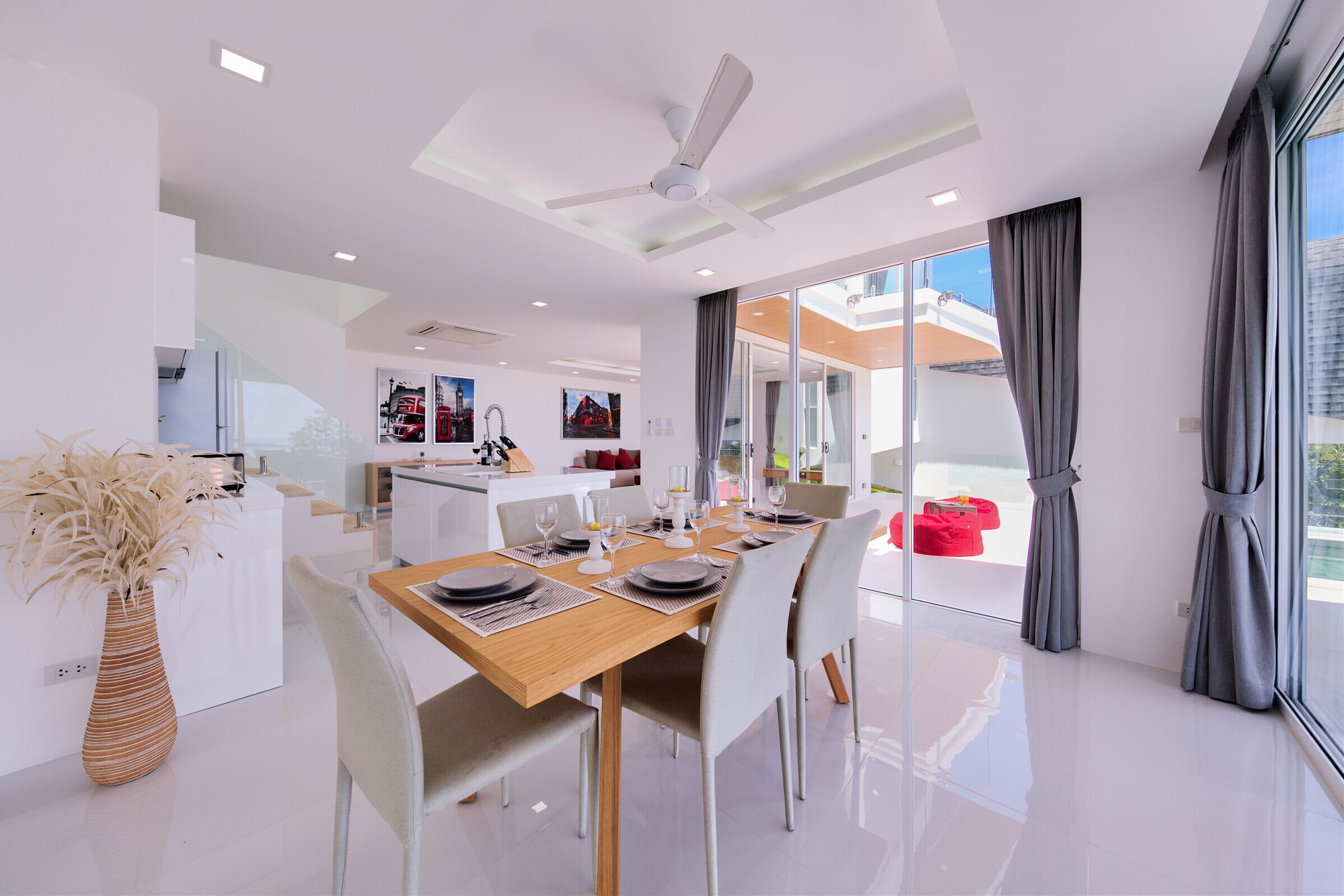 A modern dining area with a wooden table set for four, surrounded by white chairs. A vase with dried plants is on the left, and a ceiling fan is visible above.
