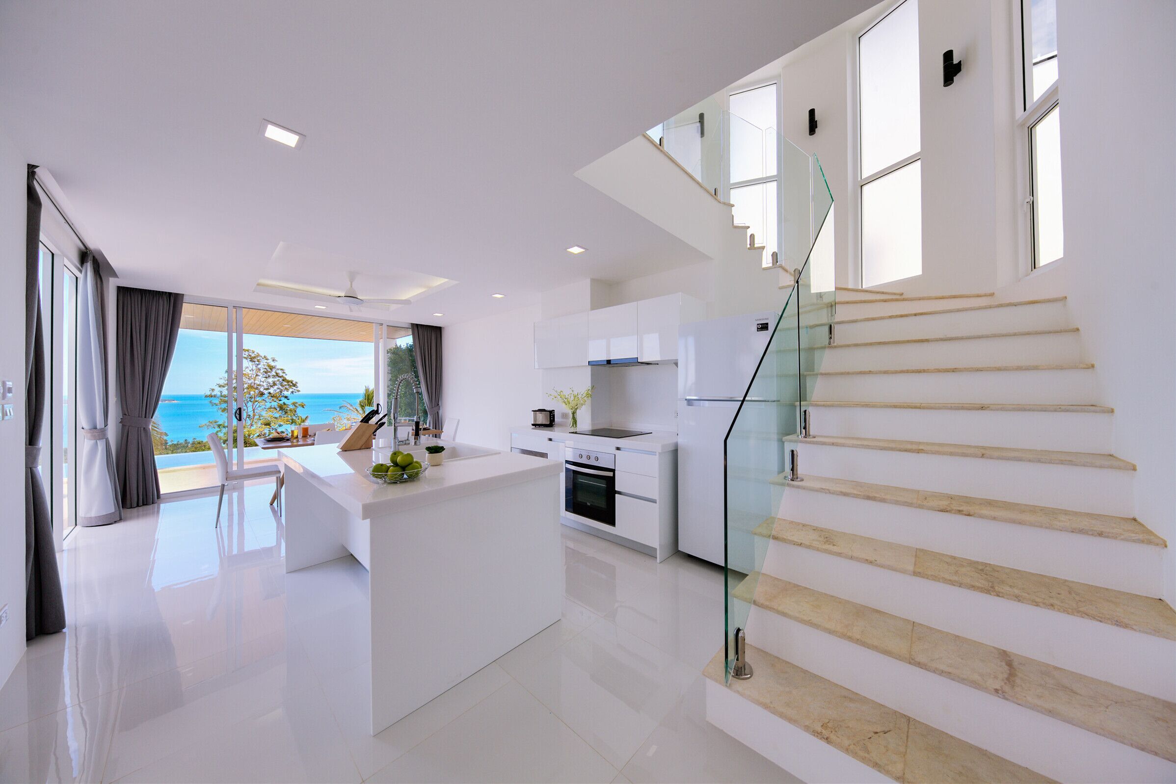 Una cocina moderna con una isla central, una escalera de cristal y vistas al océano.