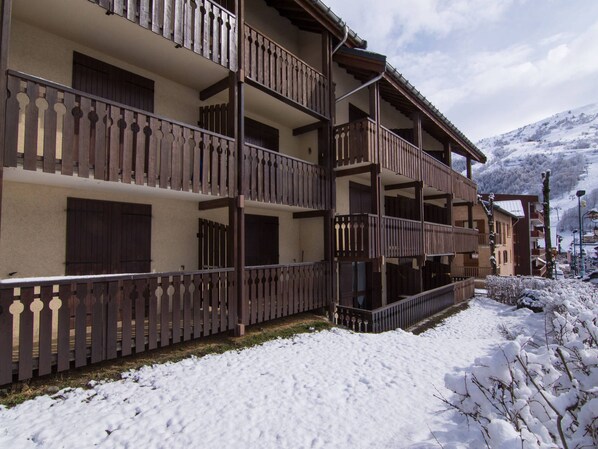 A multi-story building with balconies, a snow-covered ground, and a mountainous backdrop.