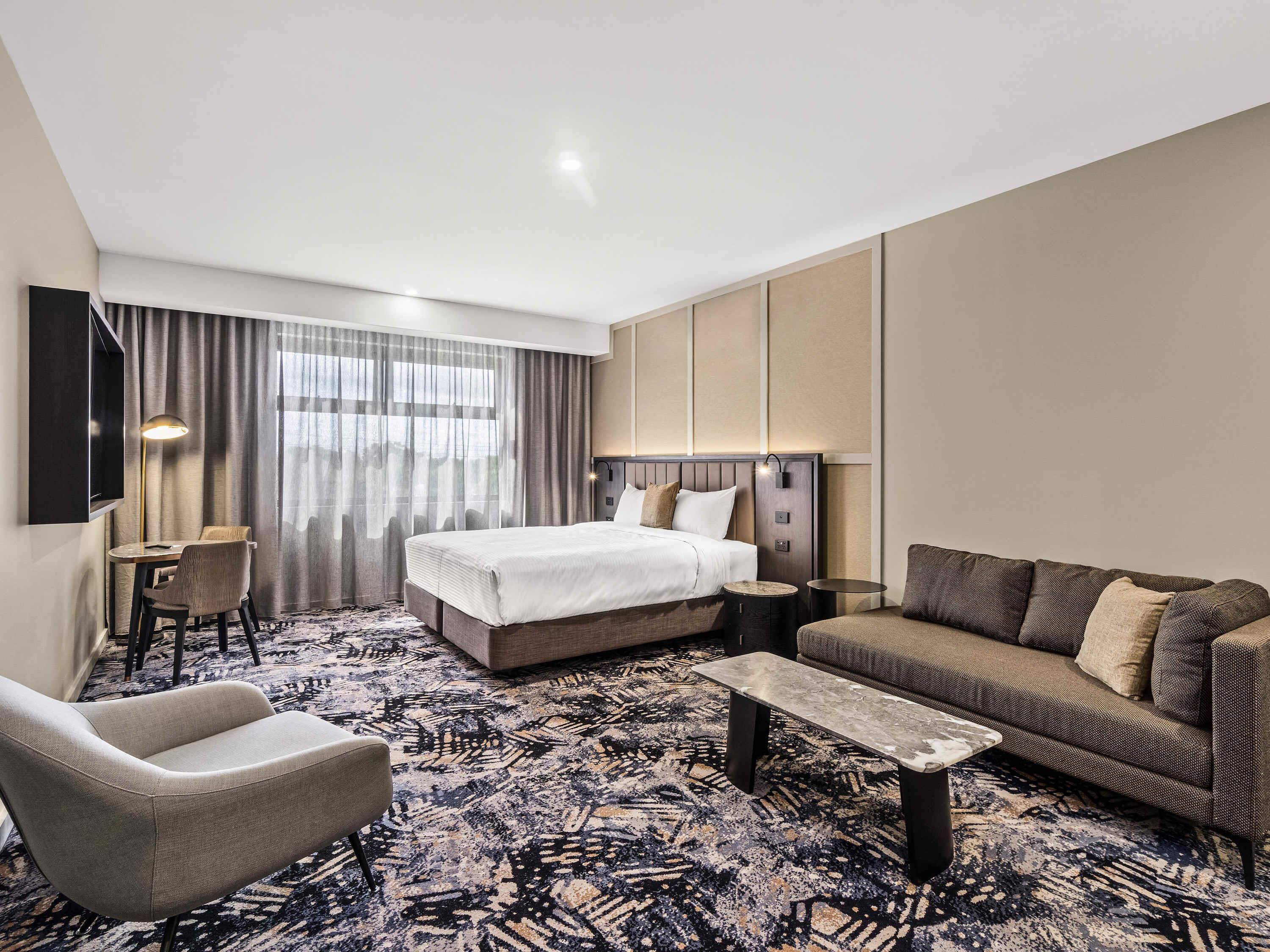 A hotel room with a bed, a sofa, a desk with a chair, a coffee table, and a TV mounted on the wall.