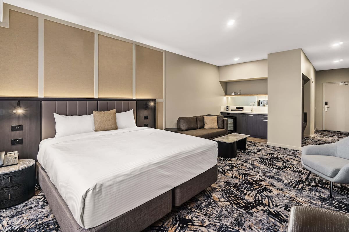 A hotel room with a large bed, a sofa, a small table, a kitchenette, and a bathroom.