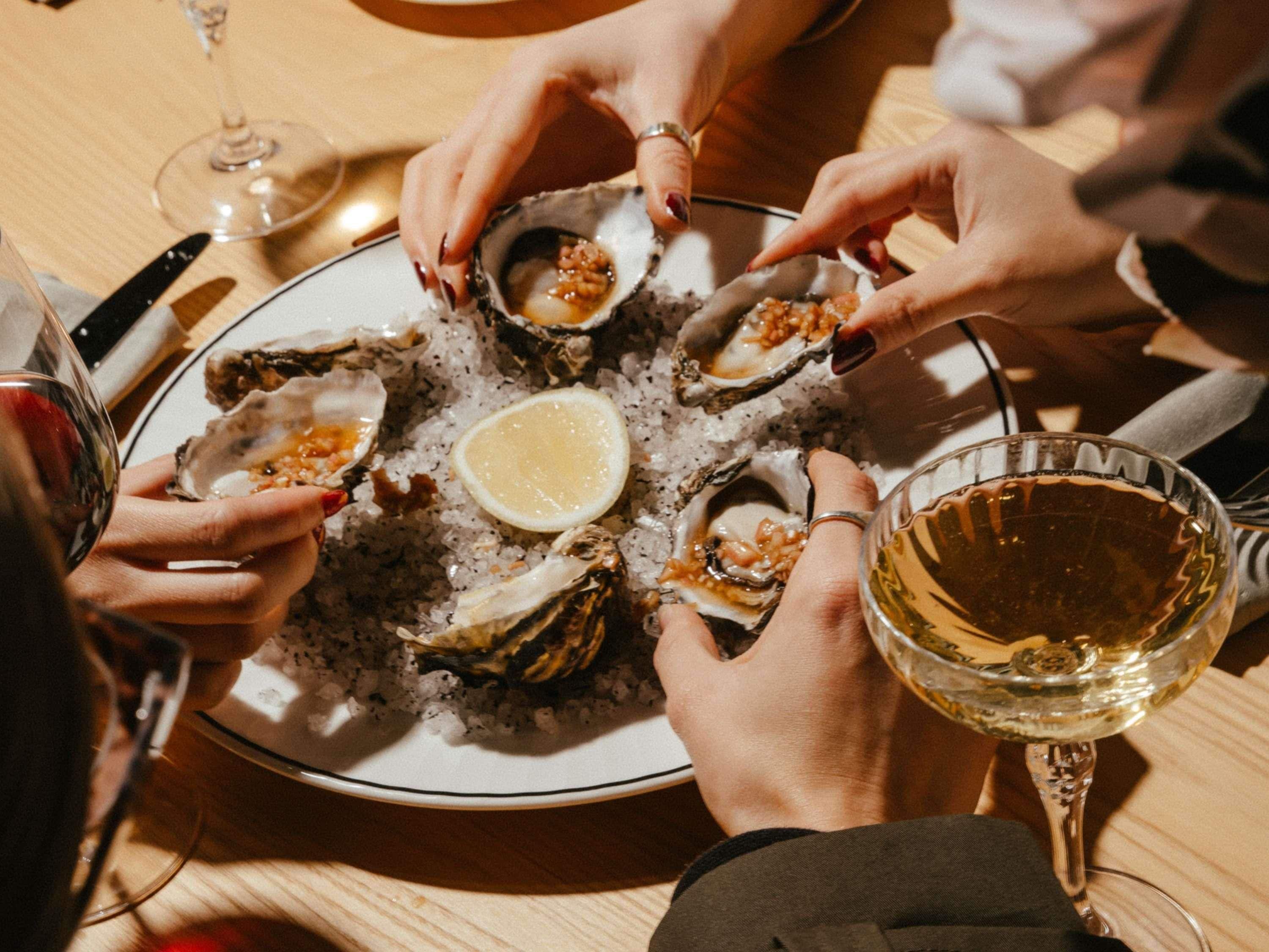 Un groupe de personnes est réuni autour d’une table, dégustant des huîtres présentées dans une assiette, accompagnées d’un verre de vin blanc.