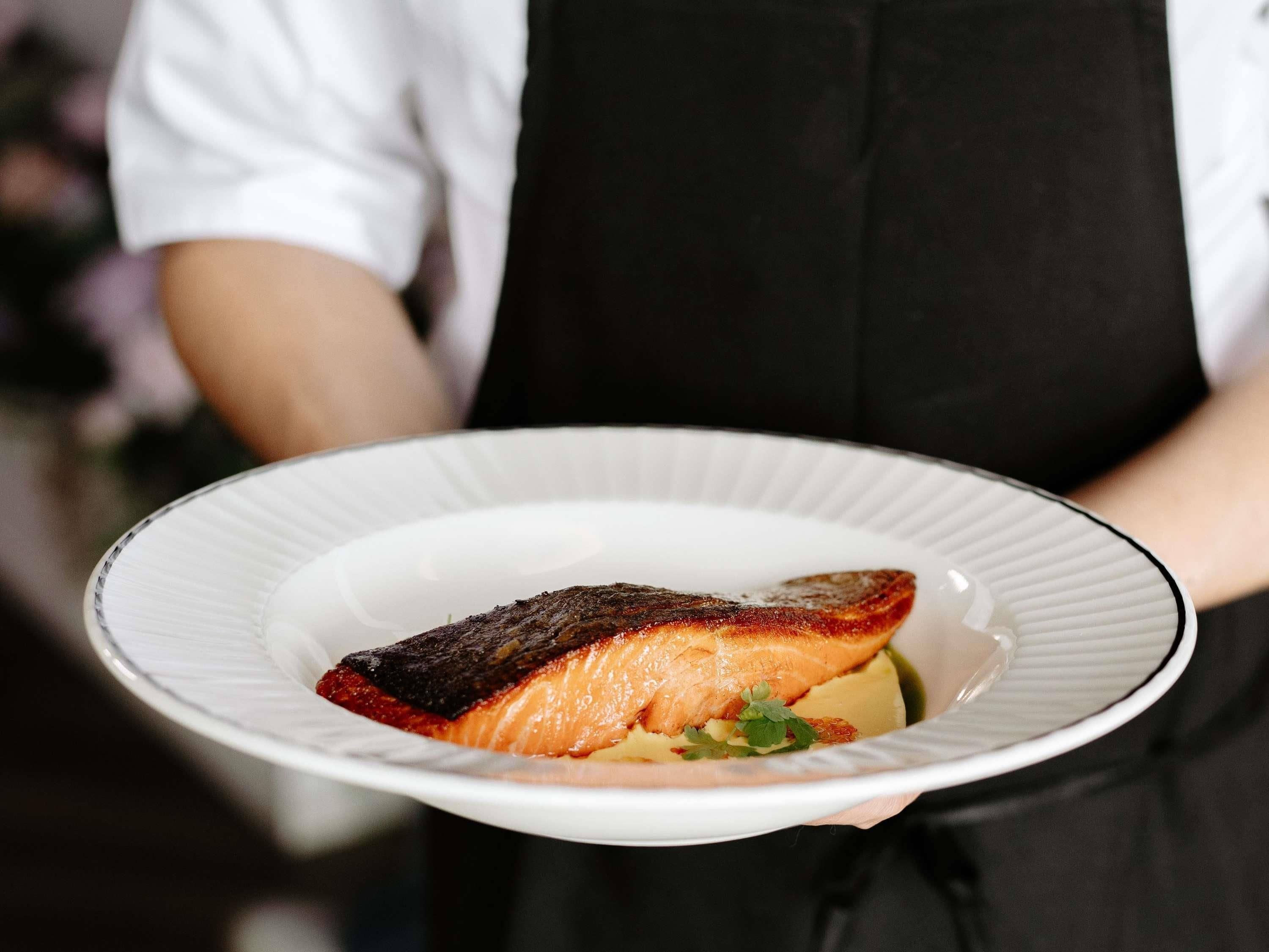 A person holding a white plate with a piece of salmon and some garnish.