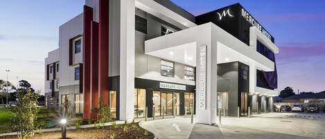 A modern hotel with a prominent entrance, a landscaped garden area, and a clear sky.