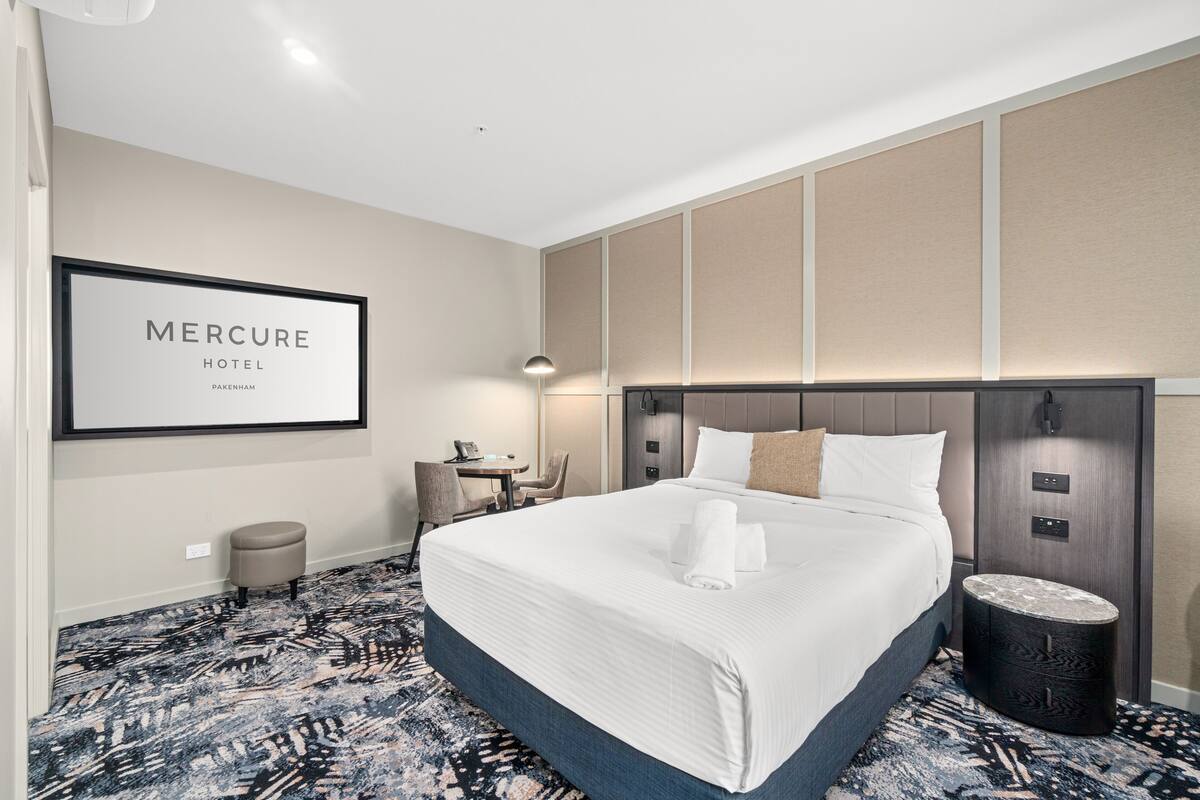 Habitación de hotel con una cama grande, un escritorio, una silla, un taburete pequeño y un cartel enmarcado en la pared.
