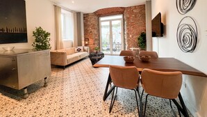 A modern living room with a sofa, dining table, and chairs. There is a stone wall and a large window.