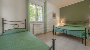 A room with two beds, a window with curtains, a radiator, and a wall-mounted lamp. - LA PALAZZINA NEL VERDE - PALAZZINA NEL VERDE N° 2 (Campo nell'Elba)