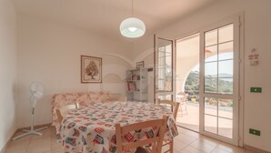 A dining area with a table covered in a tablecloth with a pattern, surrounded by wooden chairs. There is a standing fan, a bookshelf, and a door leading to an outdoor area with a view of the landscape. - LA PALAZZINA NEL VERDE - PALAZZINA NEL VERDE N° 2 (Campo nell'Elba)