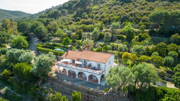 A white villa with a red-tiled roof, surrounded by lush greenery and a stone wall.