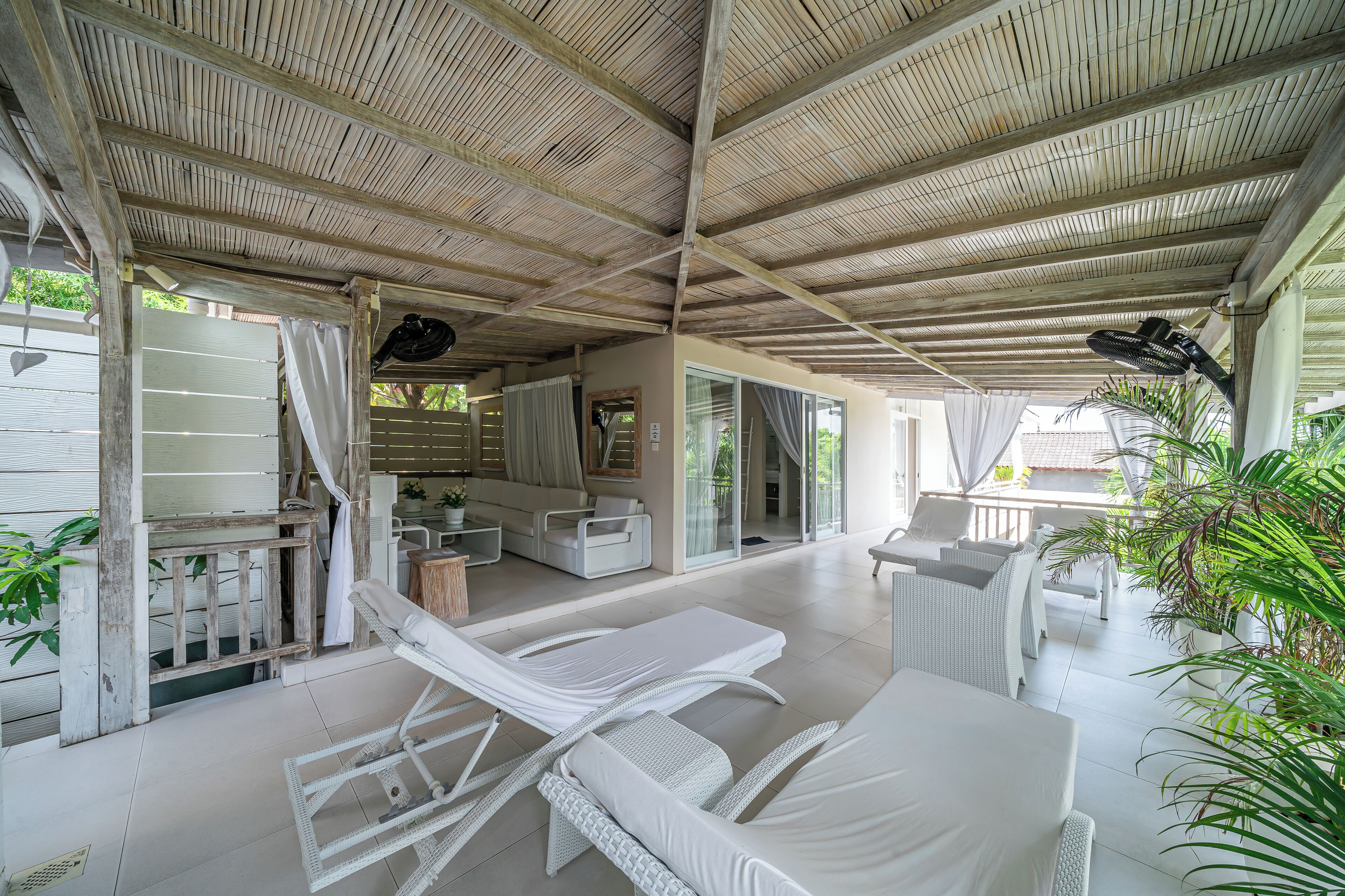 Ein überdachter Außenbereich mit Sitzgelegenheiten aus weißem Rattan, einer Decke mit Bambusleisten und Blick auf das Nachbargebäude.