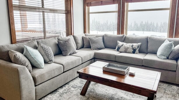 A living room with a grey sectional sofa, a wooden coffee table, and large windows with blinds. - Luxurious TRUE ski in/out chalet in Happy Valley with gorgeous views! (Beaverdell)