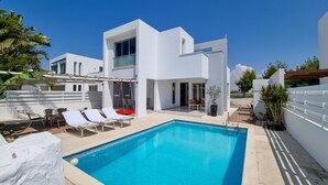 Un espace moderne aménagé au bord d’une piscine, avec une piscine rectangulaire, des murs blancs et un ciel d’un bleu limpide.