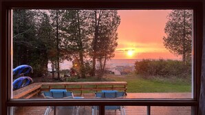 Vista al atardecer desde un balcón con banco y sillas de madera, con vista a un bosque y una masa de agua.