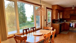 Una mesa de comedor de madera con sillas, una lámpara colgante y vistas al exterior a través de ventanas amplias.