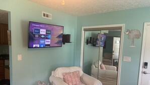 Un soggiorno con una TV a schermo piatto montata a parete che mostra diverse piattaforme di streaming. Una poltrona beige con cuscini decorati è posizionata di fronte alla TV. A sinistra, un elemento decorativo a forma di albero è posto sul pavimento.