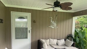 Un portico coperto con una porta bianca, un divano in vimini marrone con cuscini, un tavolino in vetro e una ventola a soffitto.