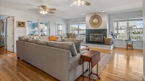 Ein geräumiges Wohnzimmer mit einem großen grauen Sofa, zwei Deckenventilatoren, einem Kamin und Blick ins Freie.
