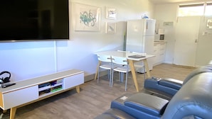 A modern living room with a flat-screen TV, a white desk with a chair, a sofa, and a refrigerator. - Getaway Villas Unit 38-7 - comfort on a budget (EXMOUTH)