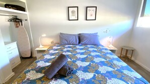 A bedroom with a bed, two pillows, a bedside table with a lamp, a nightstand, a small round table, and a wardrobe with a hanging rack. - Getaway Villas Unit 38-7 - comfort on a budget (EXMOUTH)