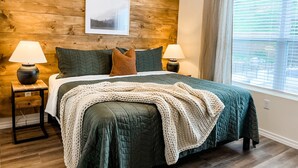 Ein Schlafzimmer mit Holz-Kopfteil, einem Bett mit grüner Bettwäsche, zwei Nachttischlampen, einem gerahmten Bild an der Wand und einem Fenster mit Jalousien.