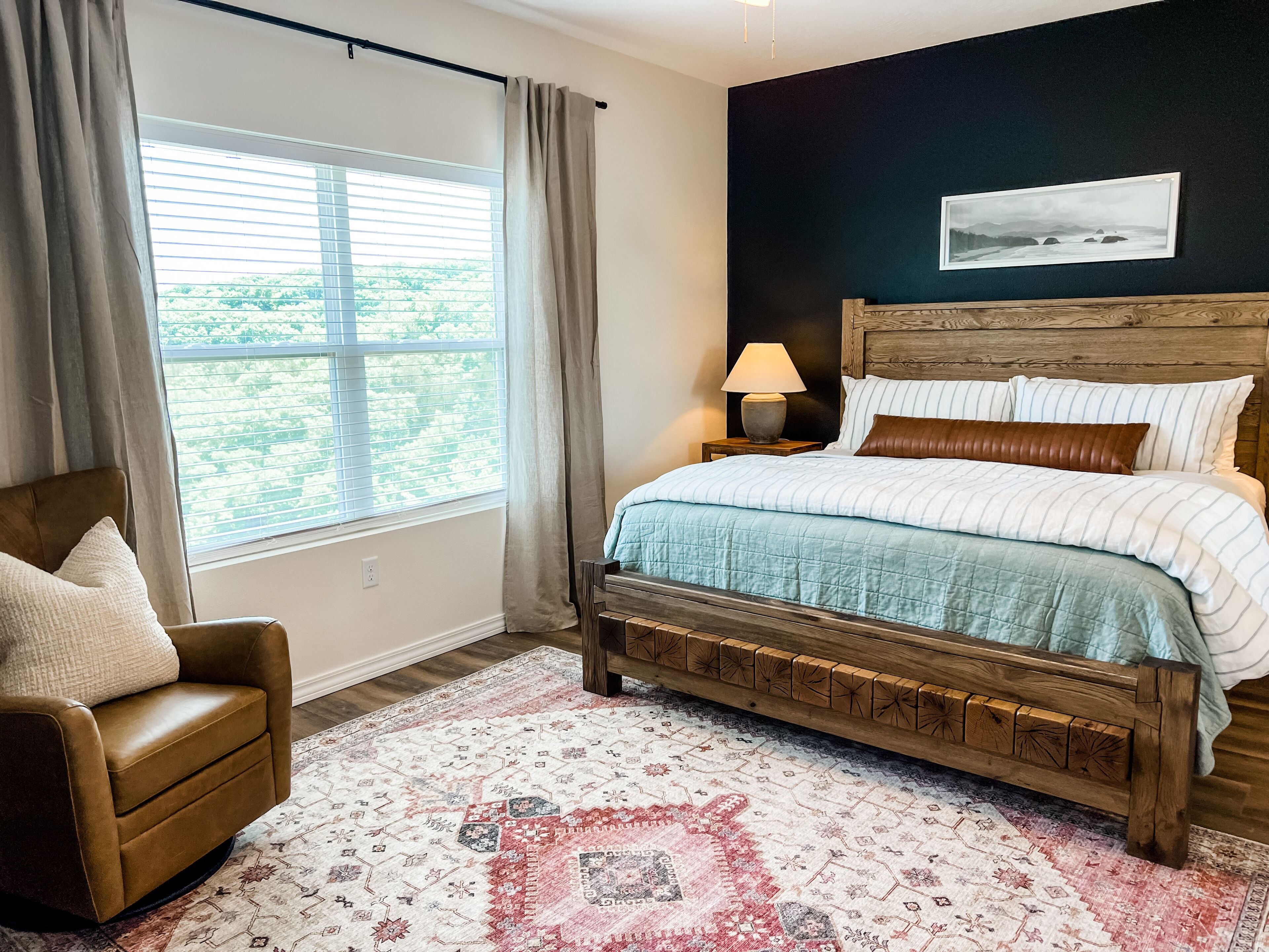A bedroom with a bed, a chair, a nightstand with a lamp, a window with curtains, and a wall-mounted picture.