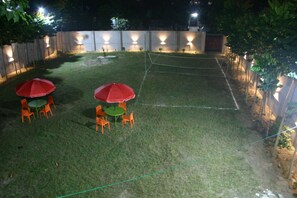 Un'area esterna ben illuminata con prato, due tavoli con sedie e due ombrelloni rossi.