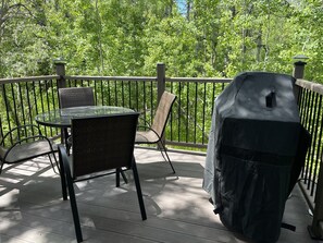 Une terrasse en bois avec une table et des chaises en verre, un barbecue et une rambarde donnant sur une zone boisée.