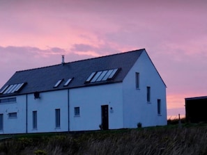 Un bâtiment blanc, doté d’un toit en pente et de nombreuses fenêtres, dont des fenêtres de toit, se dressant sur fond de ciel crépusculaire.