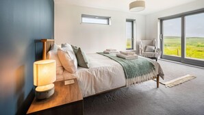 Une chambre dotée d’un grand lit, d’une table de chevet avec une lampe, d’un fauteuil et offrant une vue sur la campagne grâce à de grandes fenêtres.
